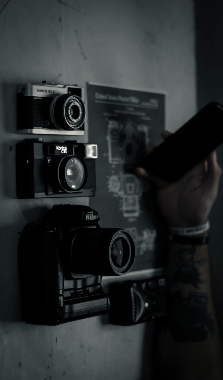 High angle of crop anonymous tattooed male browsing smartphone while sitting at table with professional photo cameras